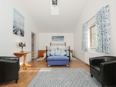 A bedroom with a metal bed frame a blue upholstered bench two black armchairs a wooden table with a lamp patterned curtains and a skylight at Lambley View in Bude