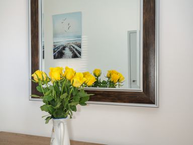 A bouquet of yellow roses in a white vase on a wooden surface with a large mirror and a beach pathway photo on the wall at Lambley View in Bude