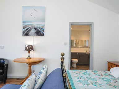 A bedroom with a bed with a blue quilt and patterned pillows a wooden table with a lamp a black armchair a painting of a beach scene and an open door to a bathroom at Lambley View in Bude