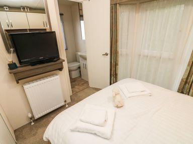 A bedroom with a bed folded towels and a teddy bear a television mounted above a radiator and an open door to a bathroom at 3 Yealands Carnforth