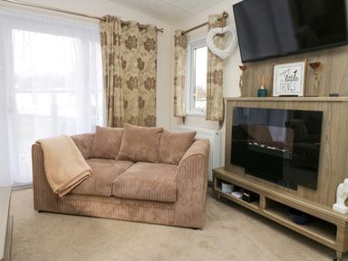 A living room with a brown sofa beige curtains a mounted television and a wooden cabinet with decorative items at 3 Yealands Carnforth