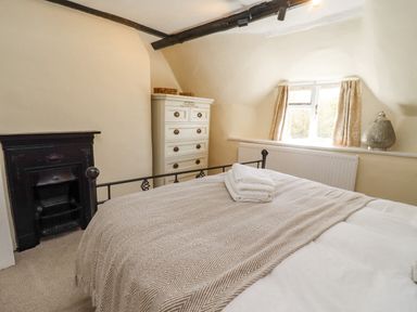 A bedroom with a bed covered by a blanket a white chest of drawers a small window with curtains and a black fireplace at 5 Packhorse in Purton