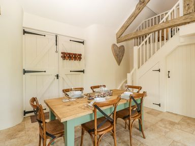 A dining area with a wooden table set for six, wooden chairs, a staircase, and double doors at The Hayloft in Stockland Honiton