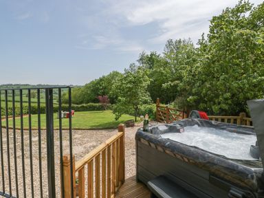 An outdoor hot tub on a wooden deck with a bottle and glass overlooking a garden and green fields at The Hayloft in Stockland Honiton
