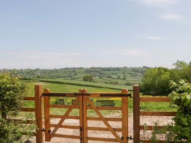 A wooden gate opening to a grassy field with hills and trees in the background at The Hayloft in Stockland Honiton