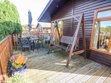 An outdoor wooden deck with a swing, table and chairs, potted plants, and wooden railing at Mallard Lodge in Carnforth