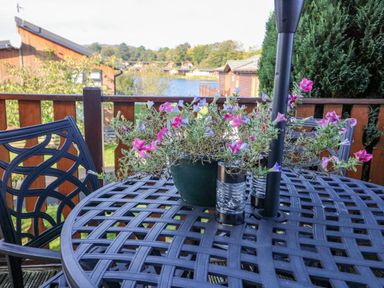 A patio table with a potted plant and two salt and pepper shakers on a balcony overlooking a lake and wooden cabins at Mallard Lodge in Carnforth