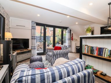 A living room with striped sofas a bookcase and large windows showing a patio at Tower End Cottage in Melton