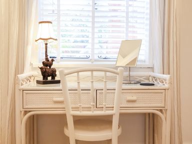 A white desk with drawers a chair a decorative lamp and a mirror in front of a window with blinds at Tower End Cottage in Melton