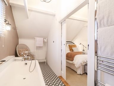 A bathroom with a bathtub and a chair next to it with a view into a bedroom with a bed and a lamp at Tower End Cottage in Melton