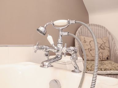 A bathtub faucet with handheld shower head and a wicker chair with cushions in the background at Tower End Cottage in Melton