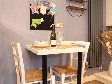 A small dining table set for two with white chairs and a bottle of wine in a room with a blue wall and a wall-mounted brochure holder at Tower End Cottage in Melton