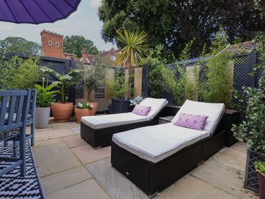 An outdoor patio with two lounge chairs with cushions and pillows surrounded by potted plants and a blue umbrella at Tower End Cottage in Melton