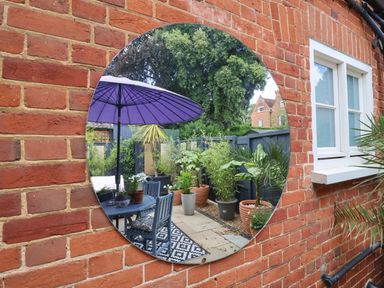 A round mirror mounted on an exterior brick wall reflecting a patio with a table, chairs, purple umbrella, potted plants, and dark fence at Tower End Cottage in Melton