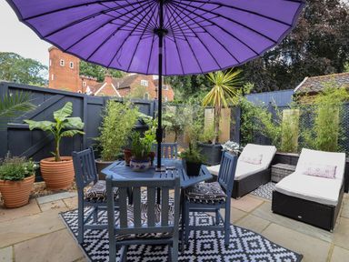 An outdoor patio with a purple umbrella above a blue table and chairs surrounded by various potted plants and two white cushioned lounge chairs at Tower End Cottage in Melton
