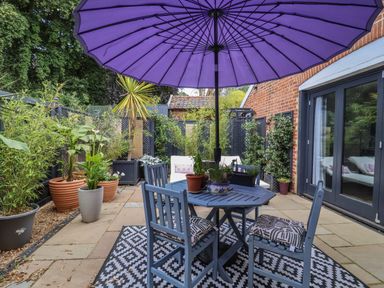 An outdoor patio with a blue table and chairs under a purple umbrella surrounded by potted plants at Tower End Cottage in Melton