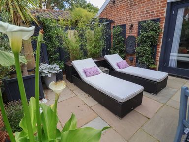 A patio area with two wicker chaise lounges with cushions and pillows surrounded by plants at Tower End Cottage in Melton
