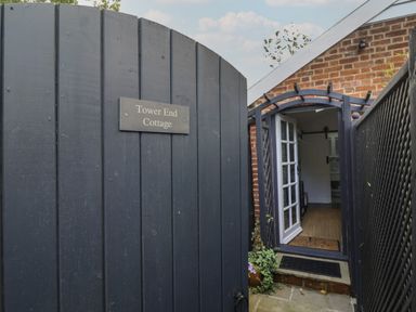 An open black wooden gate with a sign Tower End Cottage leading to a brick house entrance with an open white door and a small porch at Tower End Cottage in Melton