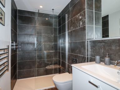 A bathroom with a walk-in shower, a white toilet, a sink with a faucet, and dark tiled walls at Porth Beach Villa in Porth near Newquay