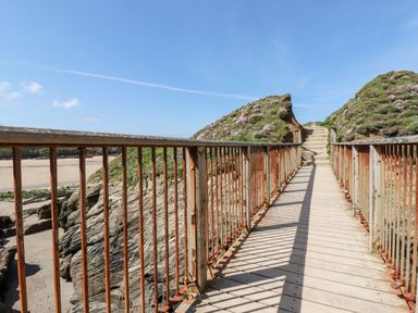 A wooden pedestrian bridge with rusty metal railings leading to stairs on a grassy hill with flowers at Porth Beach Villa in Porth near Newquay