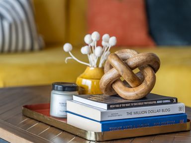 A wooden coffee table with a tray holding books a candle a yellow vase with white decorative flowers and a wooden knot sculpture with a yellow sofa and cushions in the background at Porth Beach Villa in Porth near Newquay