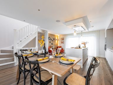 A dining area with a wooden table set with plates and yellow napkins next to a kitchen island and a seating area with orange chairs at Porth Beach Villa in Porth near Newquay
