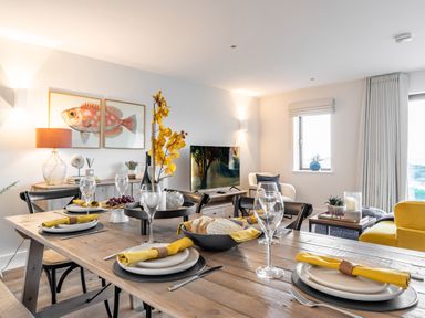 A dining area with a wooden table set with plates yellow napkins and wine glasses next to a living room with a television and chairs at Porth Beach Villa in Porth near Newquay