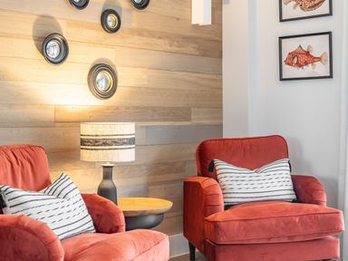 Two red armchairs with striped pillows a table lamp on a round wooden side table and wall decor with round mirrors and framed fish images at Porth Beach Villa in Porth near Newquay