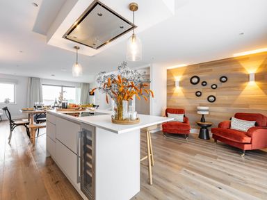 A kitchen island with flowers and bar stools next to a dining table and red armchairs in a living area at Porth Beach Villa in Porth near Newquay