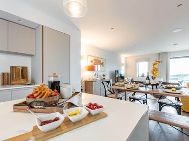 A kitchen island with croissants and condiments and a dining table set with plates and glasses in an open living space at Porth Beach Villa in Porth near Newquay
