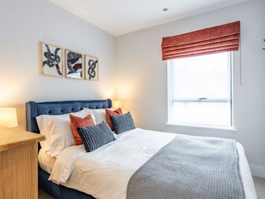 A bedroom with a bed with blue headboard white bedding pillows a wooden dresser and a window with a red blind at Porth Beach Villa in Porth near Newquay