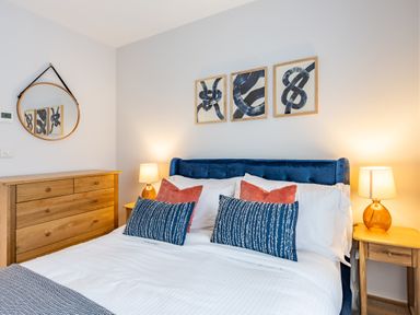 A bedroom with a blue upholstered bed framed abstract art and wooden furniture at Porth Beach Villa in Porth near Newquay