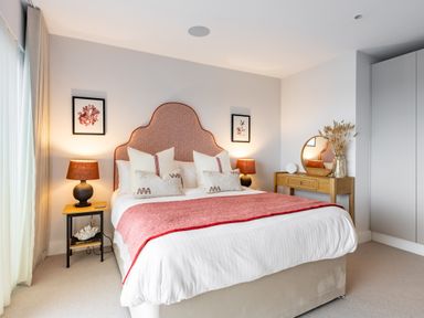 A bedroom with a large bed with coral headboard bedside tables with lamps framed pictures and a dressing table with mirror and dried flowers at Porth Beach Villa in Porth near Newquay