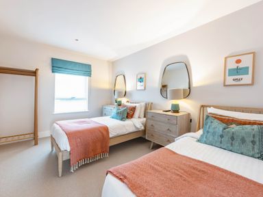A bedroom with two single beds with orange throws and decorative pillows two wooden dressers with lamps two mirrors and framed artwork at Porth Beach Villa in Porth near Newquay