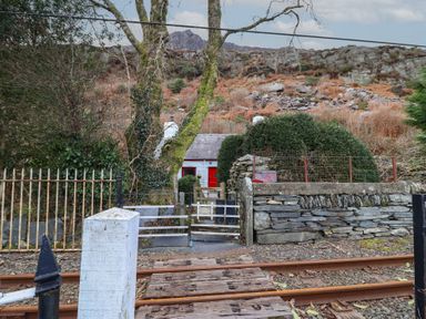 A small white cottage with a red door behind a gate near train tracks and rocky hilltop at Quarry Cottage in Blaenau Ffestiniog