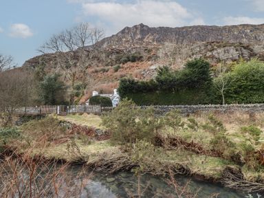 A small white cottage behind trees and hedges with a mountain in the background and a stream in the foreground at Quarry Cottage in Blaenau Ffestiniog