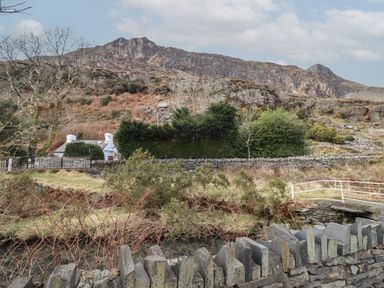 A white cottage behind trees and a stone wall with a mountain in the background at Quarry Cottage in Blaenau Ffestiniog