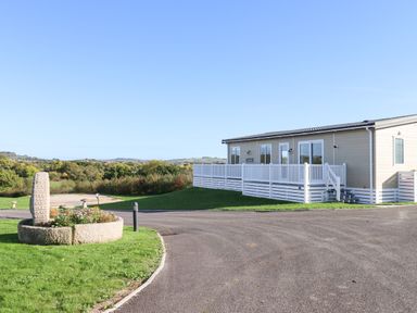 A beige single-story house with white railings on a paved road with grass and a stone flower planter in the foreground at Cherry Lodge Teigngrace near Newton Abbot