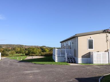 A beige mobile home with a white porch and railing near a paved road and green fields at Cherry Lodge in Teigngrace near Newton Abbot