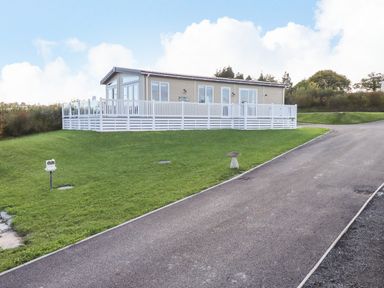 A single story mobile home with white fencing on a grassy plot next to a paved road at Cherry Lodge in Teigngrace near Newton Abbot