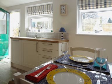 A kitchen with a dining table set for two near two windows with striped blinds and a green door at Fiddlesticks Cottage in Beaminster