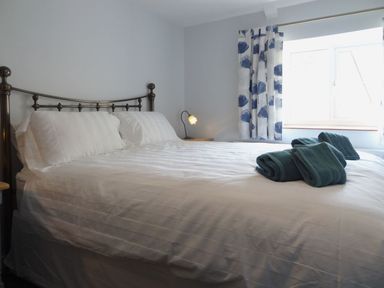 A bedroom with a bed made with white linens blue towels on the bed and a window with blue and white curtains at Fiddlesticks Cottage in Beaminster