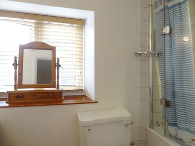 A bathroom window with wooden blinds and a wooden mirror stand above a toilet next to a bathtub with a striped shower curtain at Fiddlesticks Cottage in Beaminster