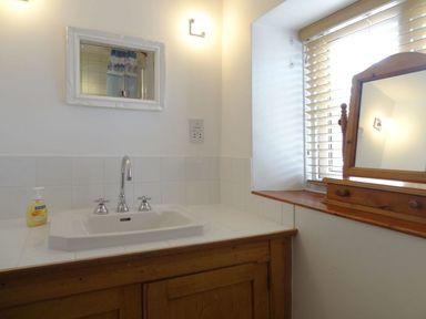 A bathroom sink with a soap dispenser a framed mirror a wooden cabinet and a wooden framed mirror on a windowsill with blinds at Fiddlesticks Cottage in Beaminster