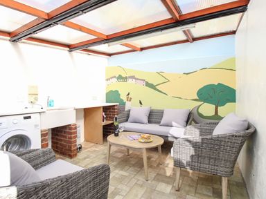 A sunroom with wicker furniture a low table a washing machine and a countryside mural on the wall at Fiddlesticks Cottage in Beaminster