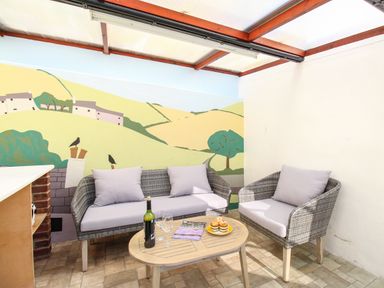 A sitting area with two grey cushioned chairs a wooden table with wine glasses a bottle and muffins and a mural of hills on the wall at Fiddlesticks Cottage in Beaminster