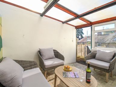 A conservatory with wicker armchairs and a sofa with cushions a wooden table with wine bottle glasses and muffins at Fiddlesticks Cottage in Beaminster
