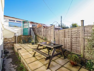A patio with a wooden picnic table set with wine and food two chairs near a green door on a raised platform fenced yard at Fiddlesticks Cottage in Beaminster