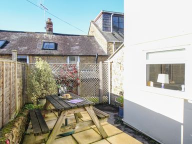 An outdoor patio with a wooden picnic table wine glasses a bottle and plants at Fiddlesticks Cottage in Beaminster