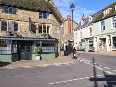 A street corner with a restaurant and shops with old stone buildings and a lamppost at Fiddlesticks Cottage in Beaminster
